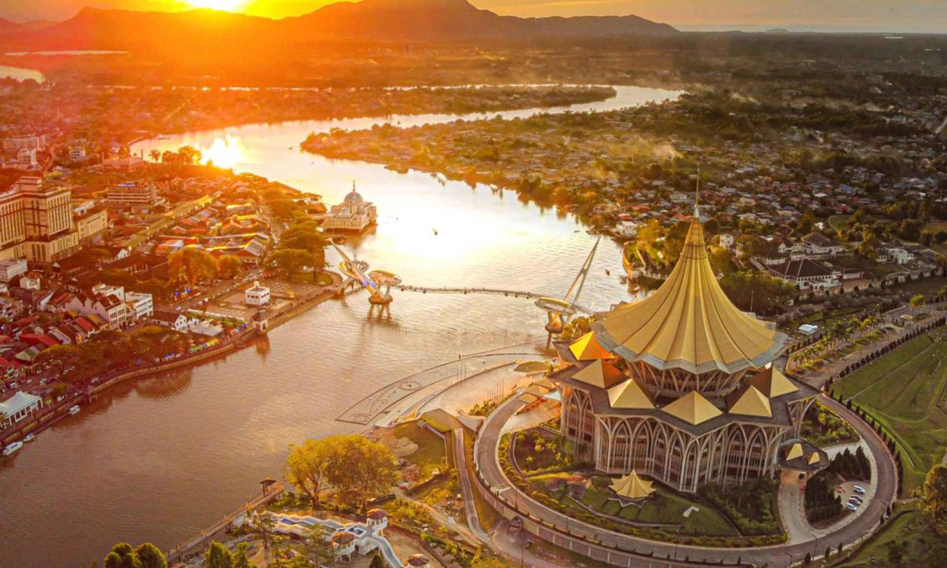 The Sarawak River winding past the gold-roofed State Assembly building in Kuching at golden hour, with Mount Santubong on the horizon.